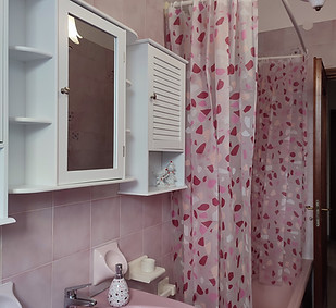 Bagno con lavabo, specchiera e mobiletti bianchi a parete, tenda doccia con fantasia di foglie rosa e lilla, piastrelle rosa sulle pareti, il tutto in un ambiente luminoso e ordinato.