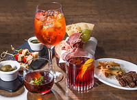 Tris di cocktail serviti su un tavolo elegante: un Aperol spritz in calice con ghiaccio, un drink scuro guarnito con frutti rossi e un cocktail con scorza d'arancia. Accanto, stuzzichini salati, olive, salumi affettati e focaccia.