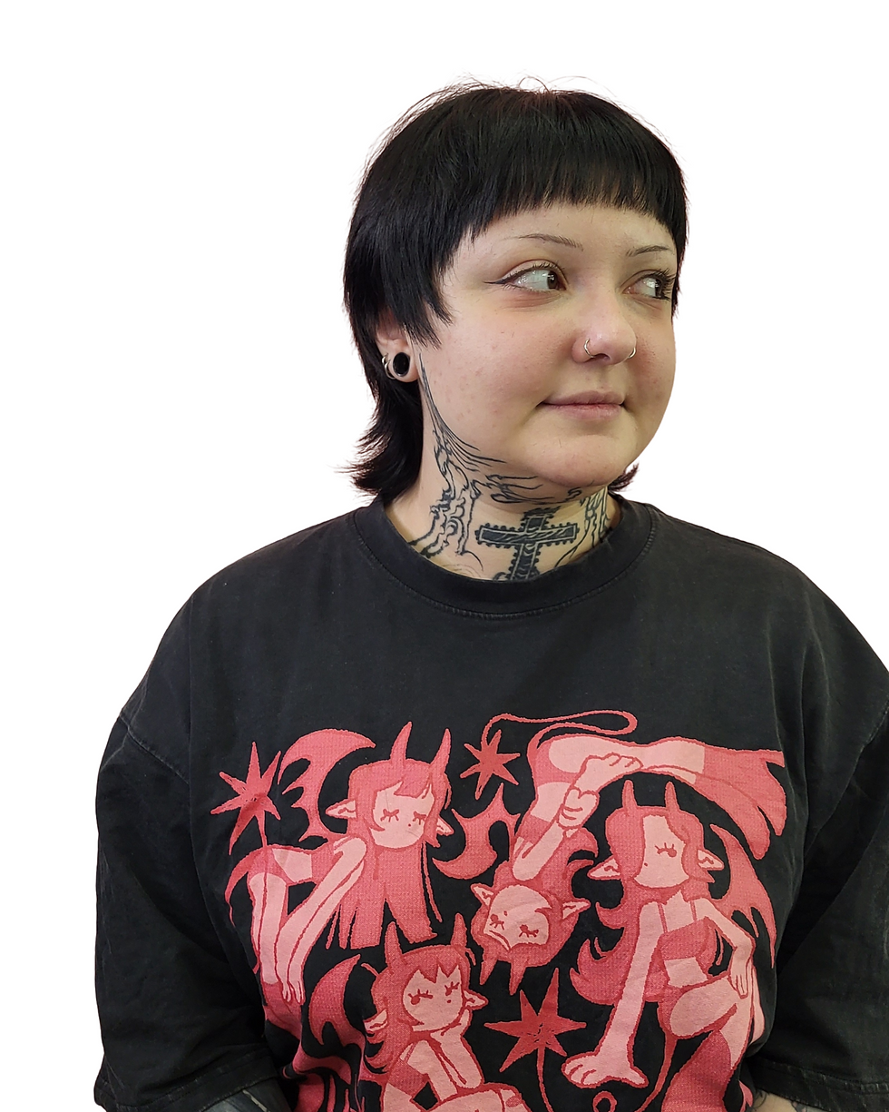 Person with black hair and facial tattoos wears a black shirt with pink cartoon demon designs. They look to the side, with a neutral expression.