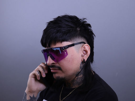 Homme coiffé d'un mulet, portant des lunettes violettes, parlant au téléphone. Fond gris, expression concentrée.