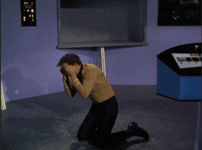 A person in a yellow shirt kneels on a spaceship floor, covering their face in apparent distress. Controls and screens are in the background.