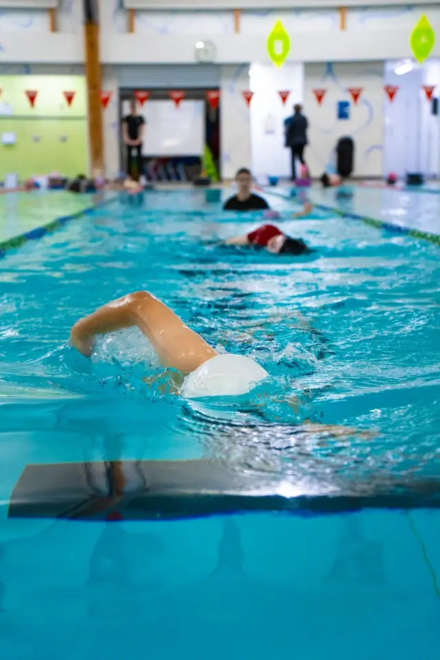 Swimmer in a white cap doing freestyle in an indoor pool, with blurred swimmers and red lane flags in the background.