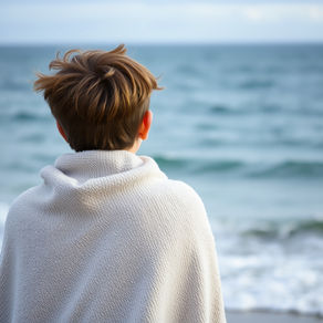 A person wrapped in a white blanket gazes at the ocean. The scene is serene, with soft waves in the background under a cloudy sky.