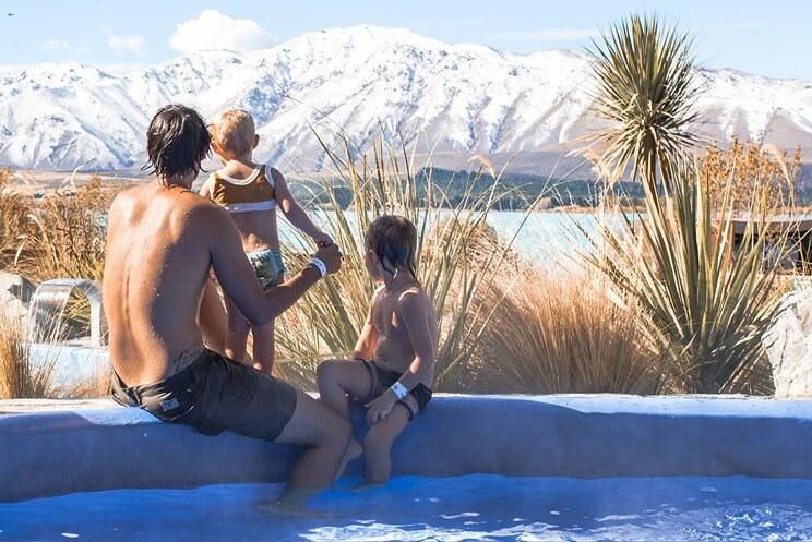 A person and two children in swimsuits enjoy a pool, with snowy mountains and a clear blue sky in the background, creating a serene mood.