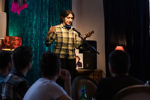Amandeep reading into the mic at queer diary at Omnibus Theatre. Amandeep is a South Asian woman with hair hair tied back wearing a grey checked shirt in front of a turquoise velvet curtain.
