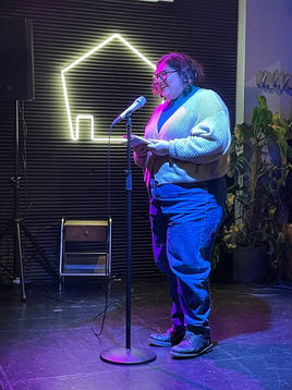 Angbeen laughing on stage under pink lights, with curly dark shoulder-length hair wearing a cream cardigan and jeans. In the background is a neon sign in the shape of a house outline.