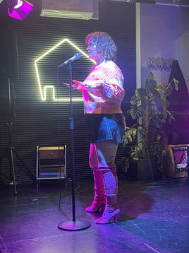 H on stage reading from a phone under pink lights, with curly shoulder-length hair wearing an orange jumper, black leather shorts, and pink knee-high boots. In the background is a neon sign in the shape of a house outline.