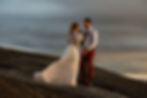A couple elopes on a mountain on their wedding day. A Woman in her white dress and knitted shawl faces away from the camera looking at a man in burnt red pants, a white button down, suspenders, and a tie. The woman's dress is blown by the wind. They are on top of a mountain with dark grey clouds in the sky.