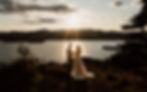 A couple embraces on top of a mountain during sunset on their elopement day. The sunflare shines on the couple with a lake and mountains in the distance. The couple has just had a hiking elopement in Lake George NY.