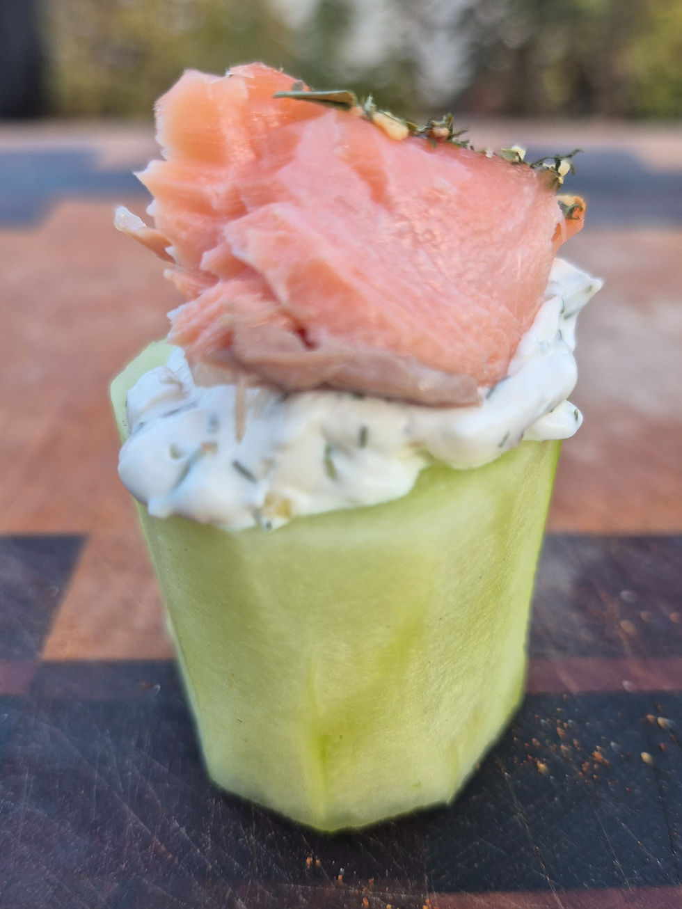 Salmon atop creamy dill on a cucumber slice, placed on a wooden cutting board, with a blurred outdoor background.