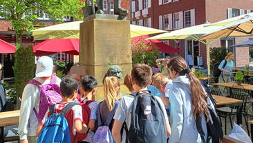 Abstecher zum Denkmal des Kiepenkerls. Die Kinder der Klasse stehen am Kiepenkerl-Denkmal. Sie schauen sich die Statur an.