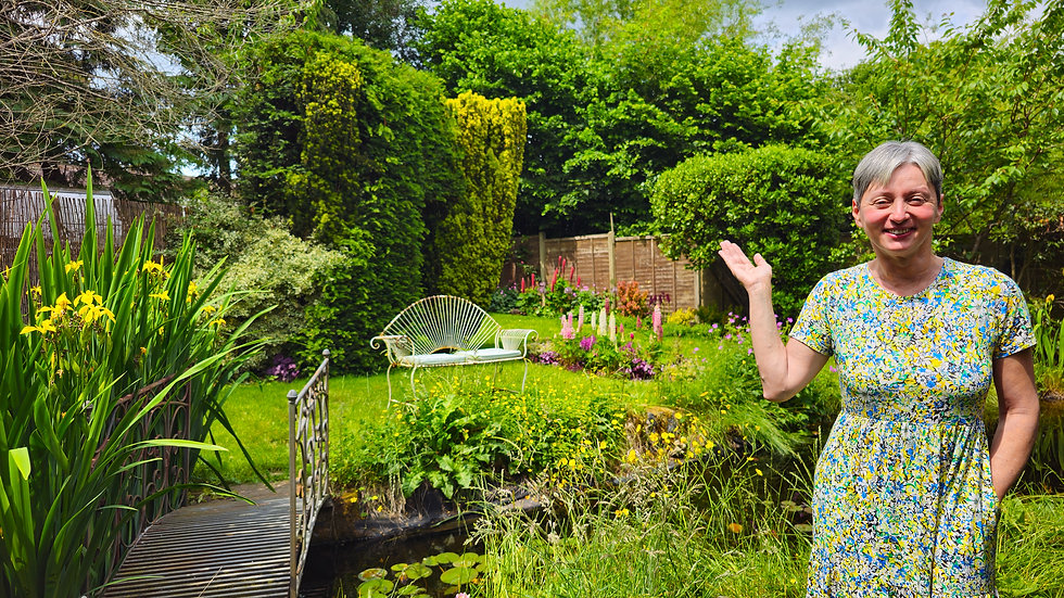 Laura with a flower dress in her cottage garden in Chawton with a lush green lawn, blooming lupins and a little metal bridge over her pond
