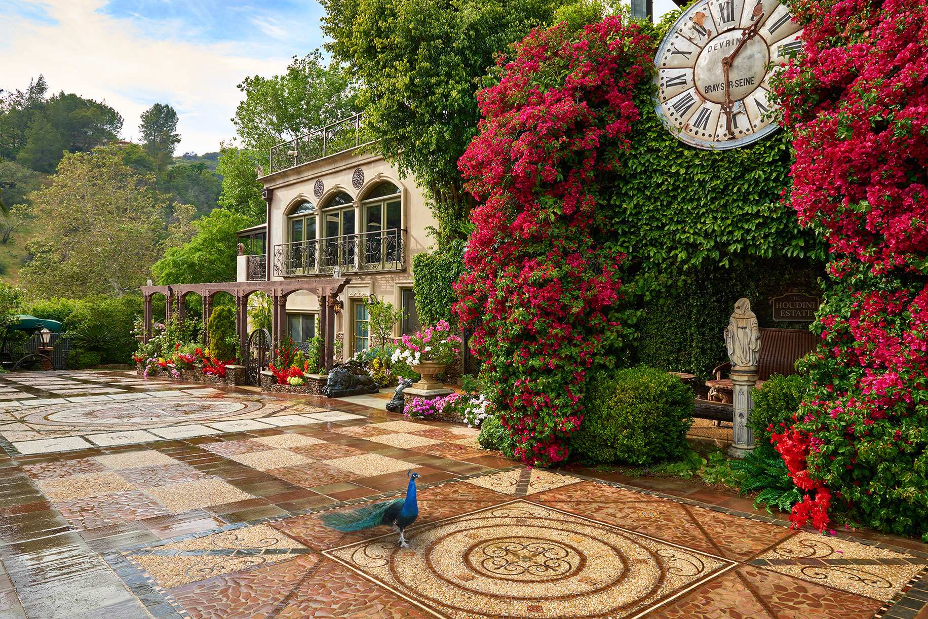 Sunlit courtyard of the Houdini Estate in Laurel Canyon, Los Angeles, with peacocks crossing the mosaic patio beneath cascading bougainvillea and the mansion facade