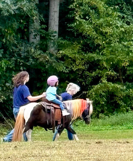 Pony with rainbow color mane being led by a woman and supported by a young man getting a ride from Haystack Homestead Moble Farm Experiences Pony & Unicorn Rides