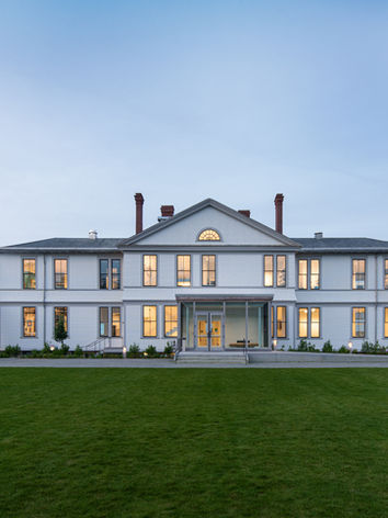 A two-story, symmetrical white building. The center portion of the building is flanked by two wings. It is sunset and the ocean is in the background.