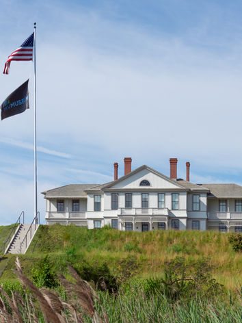 A two-story, symmetrical white building. The center portion of the building is flanked by two wings. Two flags are flying at the top of a steep staircase that leads to the lawn in front of the building.