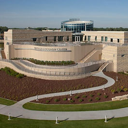 A sand colored building with a curved central section and two, taller wings. A glass tower rises above the wings at the rear. 