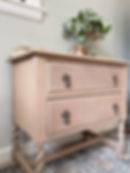 Two-drawer blush-colored chest of drawers with antique bronze hardware, flowers on top.