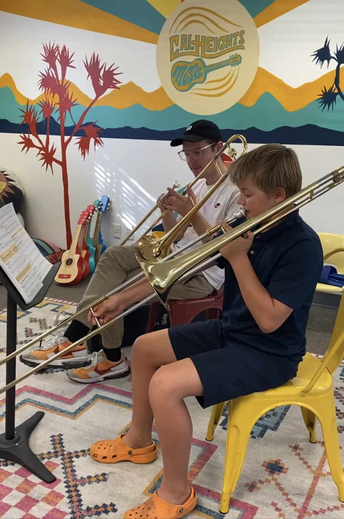 Teacher and young student in a trombone lesson in front of the Cal Heights Music branded mural