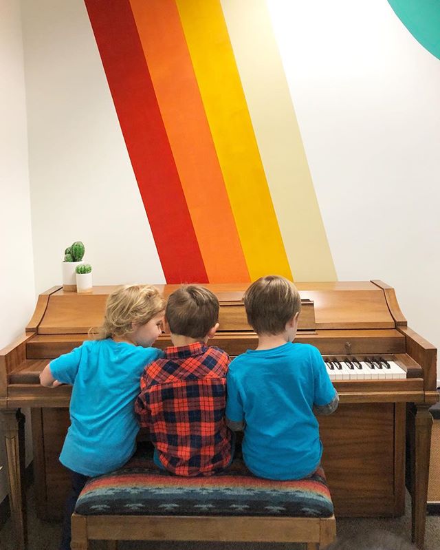 Three kids sitting together at a piano in front of the iconic Cal Heights rainbow stripe wall