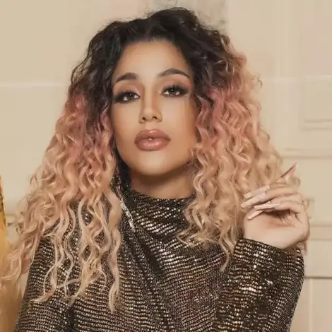 Foto de Azzy, uma mulher com cabelos cacheados em um tom de rosa claro, usando uma blusa brilhante de gola alta e manga comprida.