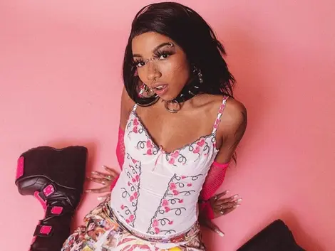 Foto de Slipmami, uma mulher sentada em um chão rosa, olhando para cima. Ela usa um corset e saia brancos, com estampa de flores rosa, e uma bota preta de salto plataforma.