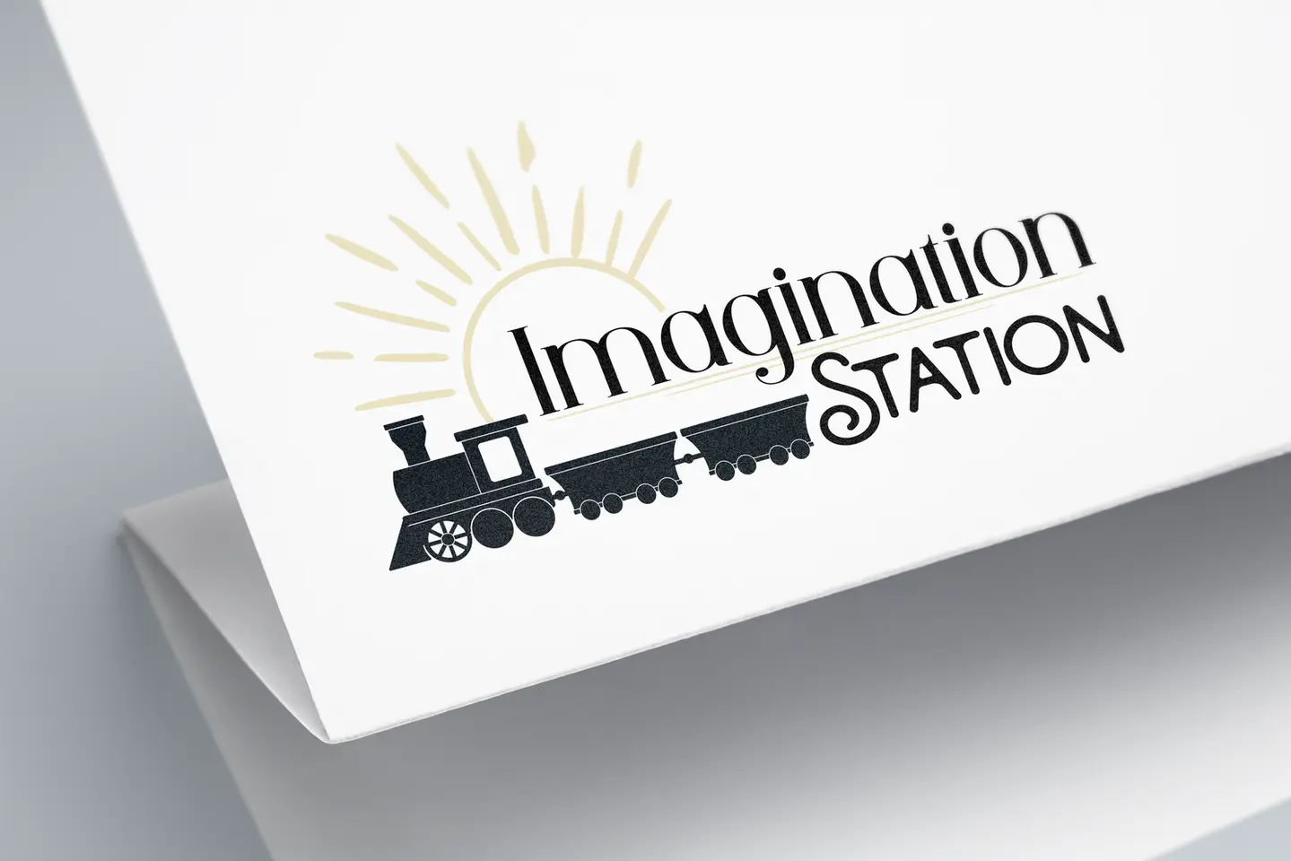 a logo design for Imagination Station that features a train outline with an inspirational sun in a pale yellow on folded pape