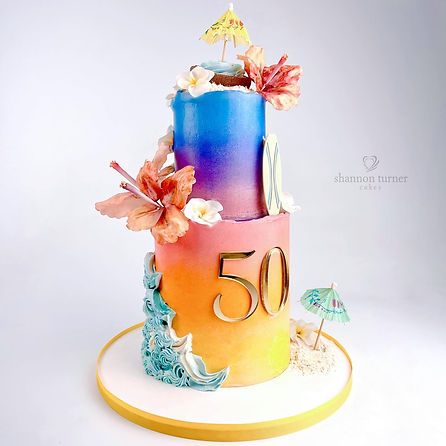 Bright and colorful two-tiered Hawaiian themed cake in bright sunset and ocean ombre faded colors, with sugar flowers, buttercream wave accents, drink umbrellas, artificial sand, a surfboard, and edible coconut details