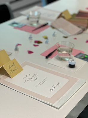 photo of a calligraphy class table with a workbook of instructions and guides, an ink pot, calligraphy pen holder, water cup, and snacks and refreshments for students to enjoy