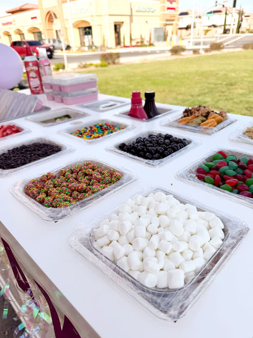 Dessert toppings bar with marshmallows, berries, and candy toppings at an outdoor event.