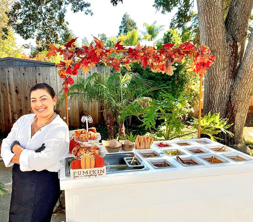 Event host standing beside a decorated mobile charcuterie cart at an outdoor gathering.