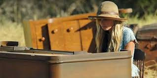 Woman in a straw hat plays an outdoor piano in a rustic yard, with a warm, focused mood.