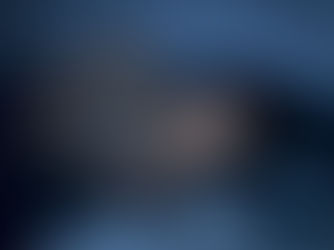 Woman lying in bed at night, looking at a phone with a focused expression. Dimly lit room with a white brick wall and a blue mug nearby.