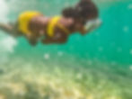Child in a yellow swimsuit swims underwater with goggles, surrounded by bubbles and rocks on the ocean floor. Bright and playful mood.