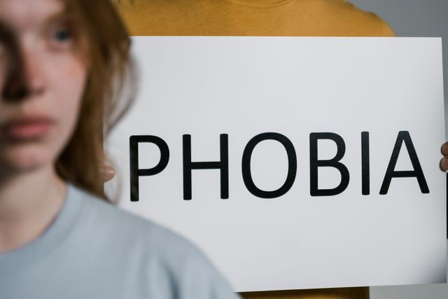 Femeie estompată în prim-plan, iar în spate o persoană ține un panou alb cu textul PHOBIA.