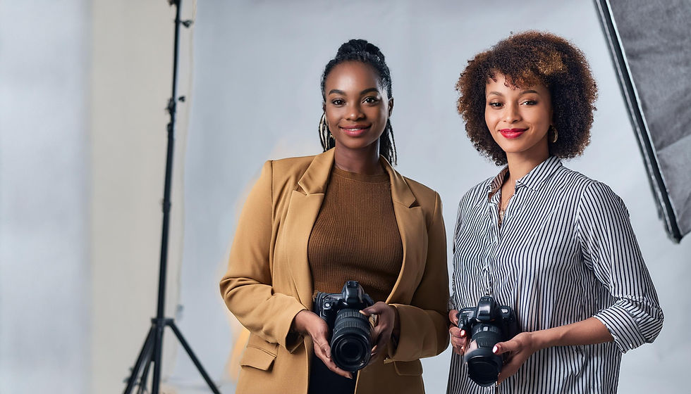 Two women smiling and holding cameras in a studio with lighting equipment, wearing a tan blazer and striped shirt, conveying confidence.
