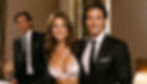 Three people in formal attire smile in a warmly lit room. One woman in a white bra; two men in black suits. Elegant and confident mood.