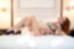 Person in black lingerie and high heels poses on white bed in a softly lit room, displaying tattoos and a relaxed expression.