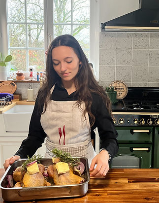 Stessie Foucart tenant un plat à gratin dans une cuisine, celui-ci contient du poulet assaisonné de beurre, huile d'olive oignons rouges et branches de thym
