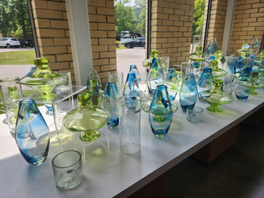 Blue and green glass vases on a white table near large windows. Sunlight casts colorful reflections. Brick wall and parked cars outside.