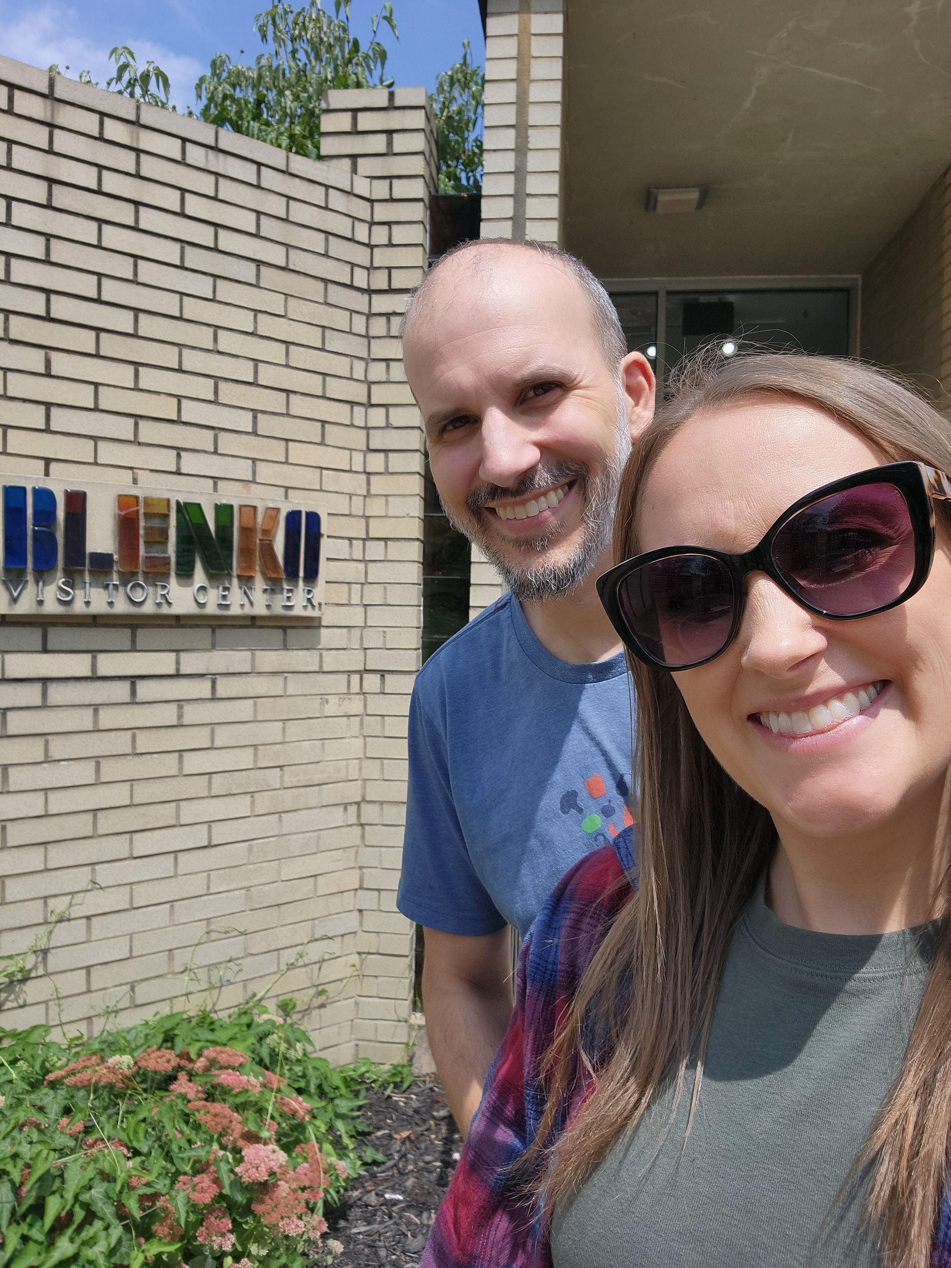 Smiling man and woman in sunglasses pose outside Blenko Visitor Center. Brick wall and green plants in the background. Bright sunny day.