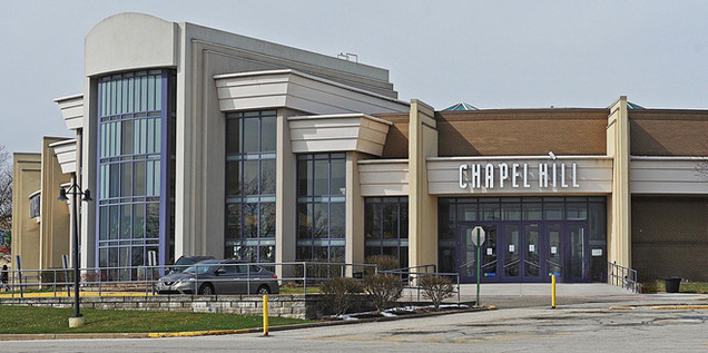 Modern building with tall glass windows, beige facade, and "Chapel Hill" sign. A parked car and a barren landscape in the foreground.