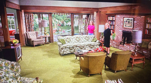 Two women stand in a vintage living room with floral furniture and brick fireplace. Large windows show a garden. Warm, retro ambiance.