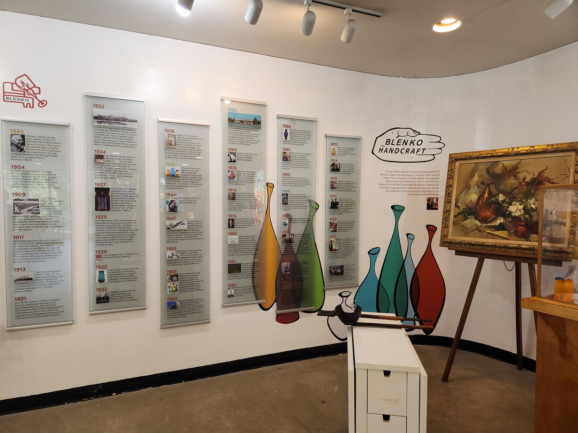Museum display with timeline panels on a wall, labeled "BLENKO HANDCRAFT." Colorful glass vase illustrations and a framed painting on an easel.