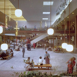 People relax and socialize in a spacious, well-lit mall with wooden accents and hanging globe lights. A sense of calm and community prevails.