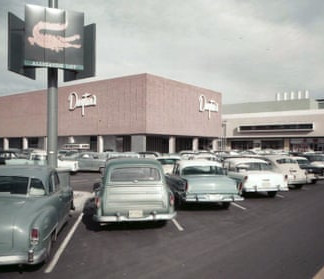 Vintage cars parked outside a brick department store with a large "Dayton's" sign. Overcast sky and a retro feel.