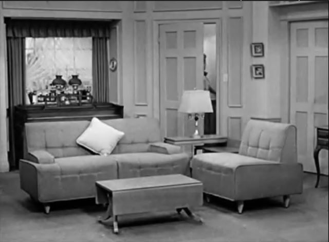 Vintage black-and-white living room with a sofa, armchair, and coffee table. A lamp is on a side table; a cabinet is visible in the background.