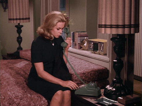 Woman in black dress on a patterned sofa, holding a green rotary phone. Dimly lit room with decorative lamps and framed pictures. She looks pensive.