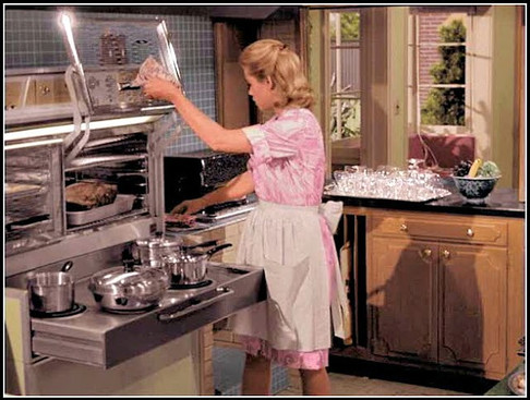 Woman in a pink dress cooking in a vintage kitchen, wearing an apron. Pots on the stove, cabinet with glassware, window view outside, daytime.