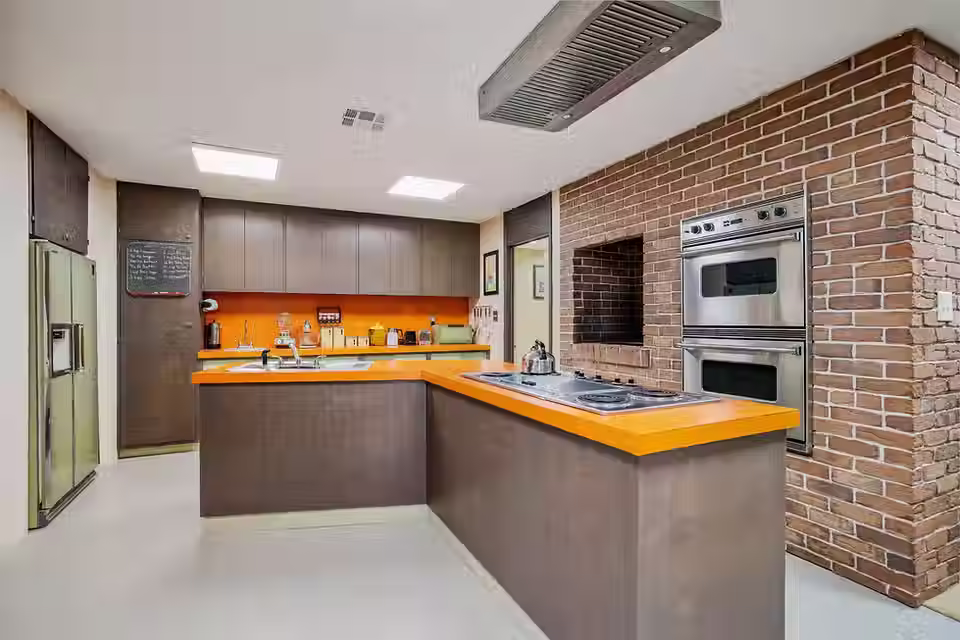 1970s kitchen with dark wood cabinets, bright orange counter tops and avocado appliances. Brick wall and stainless steel oven in the wall.Bright, warm, nostalgic